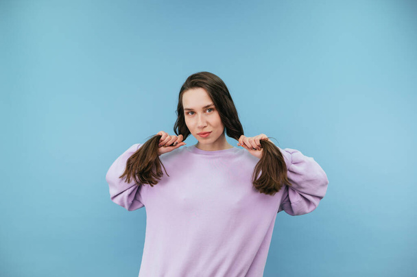 Attractive brunette woman in casual clothes holds her hands by the ponytails with her hair and poses for the camera on a blue background. - Fotoğraf, Görsel