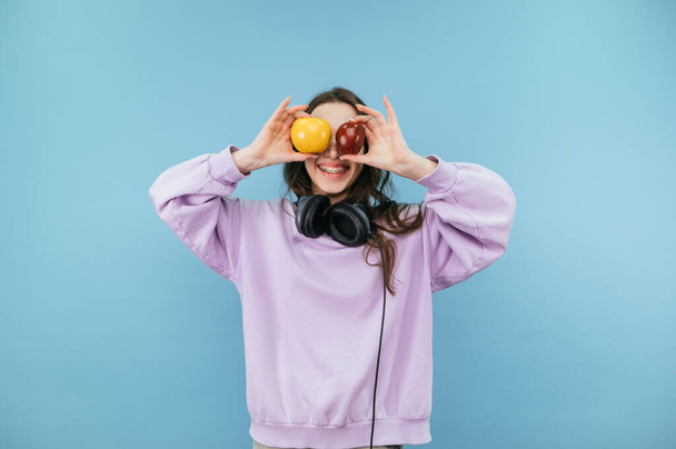 Happy girl in purple sweatshirt raised apples to eye level on blue background and smiles. - Foto, imagen