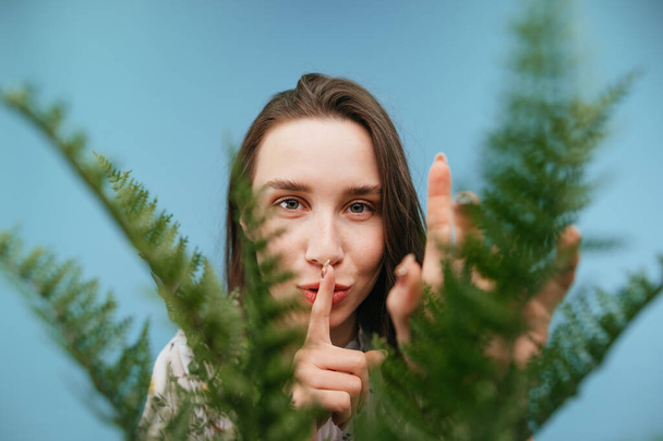 Χαριτωμένη κυρία σε μπλε φόντο με ένα πράσινο φυτό στο προσκήνιο δείχνει μια χειρονομία σιωπής και κοιτάζει την κάμερα με ένα χαμόγελο στο πρόσωπό του. - Φωτογραφία, εικόνα