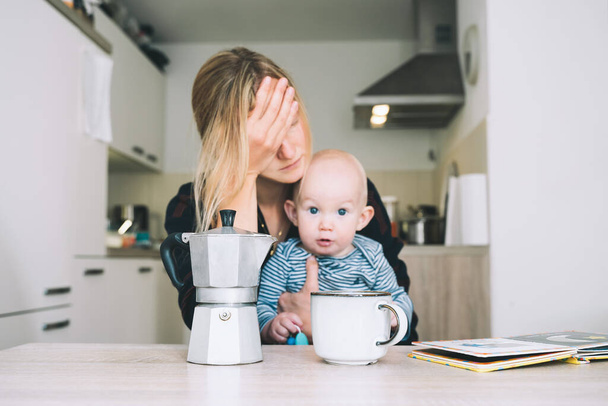 Modern young tired mom and little child after sleepless night. Exhausted woman with baby is sitting with coffee in kitchen. Life of working mother with baby. Postpartum depression on maternity leave. - Φωτογραφία, εικόνα