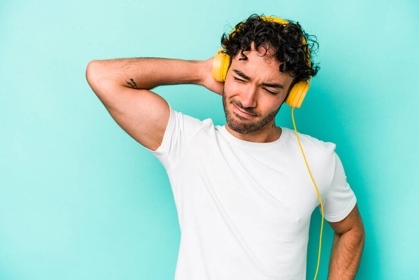Νεαρός καυκάσιος που ακούει μουσική απομονωμένος σε μπλε φόντο αγγίζοντας το πίσω μέρος του κεφαλιού, σκεπτόμενος και κάνοντας μια επιλογή. - Φωτογραφία, εικόνα