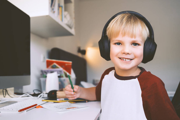 Child 7 years old is homeschooling or distance online education. Elementary school boy studying, does homework or creating at desk. Smiling kid in headphones near computer drawing or writing at home. - Zdjęcie, obraz