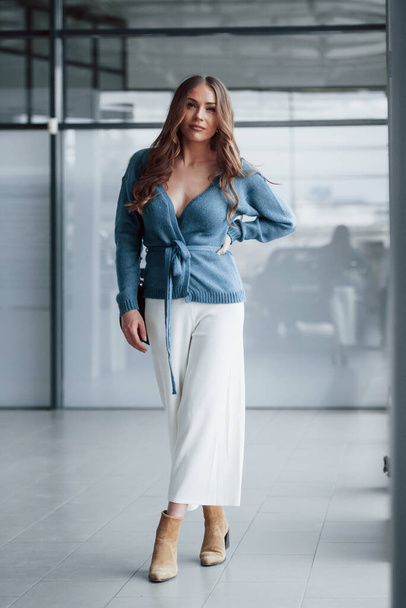 Attractive woman in blue shirt and white pants indoors posing for a camera. In airport or car shop. - Фото, зображення
