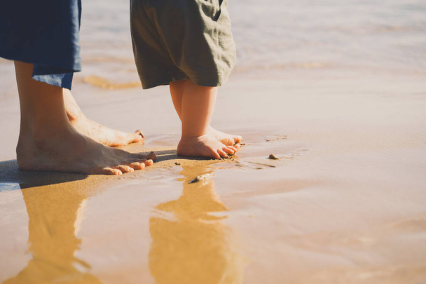 Baby's first steps. Mother and small child walking barefoot on beach sand near by ocean. Happy young family in nature. Mother and baby playing outdoors on sea beach. Close-up human feet. - Foto, immagini