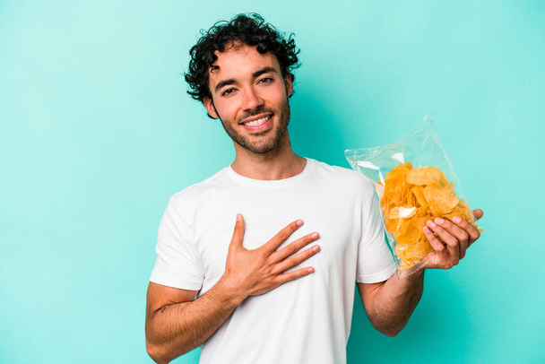 Young caucasian man holding a bag of chips isolated on blue background laughs out loudly keeping hand on chest. - Φωτογραφία, εικόνα