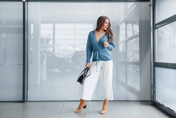 Attractive woman in blue shirt and white pants indoors posing for a camera. In airport or car shop. - Фото, изображение