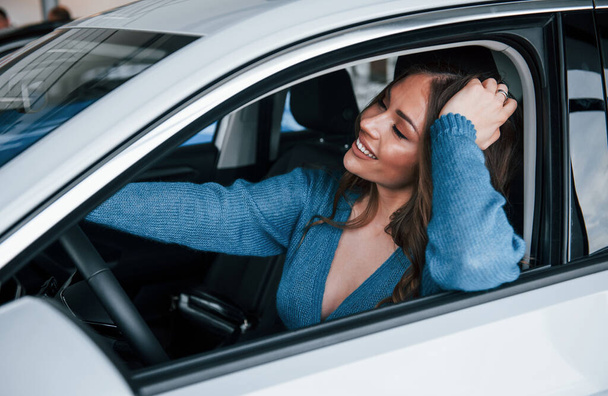 Positive woman in blue shirt sits inside of new brand new car. In auto salon or airport. - Foto, Imagem
