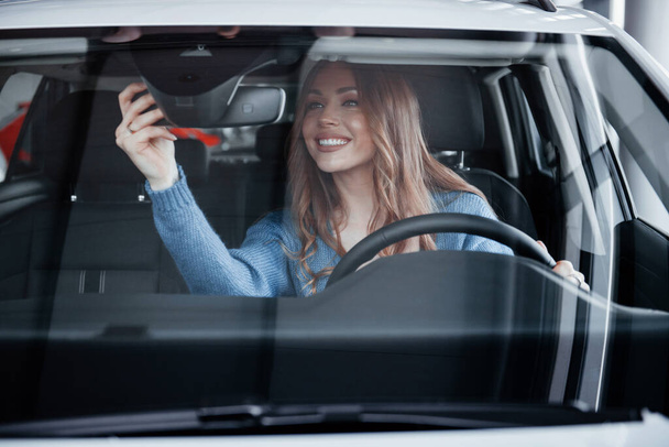 Front view of positive woman in blue shirt that sits inside of new brand new car. In auto salon or airport. - Foto, imagen