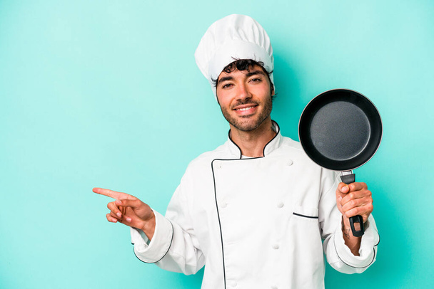 Jonge Kaukasische chef-kok met vliegende pan geïsoleerd op blauwe achtergrond glimlachend en wijzend opzij, iets tonend in de lege ruimte. - Foto, afbeelding