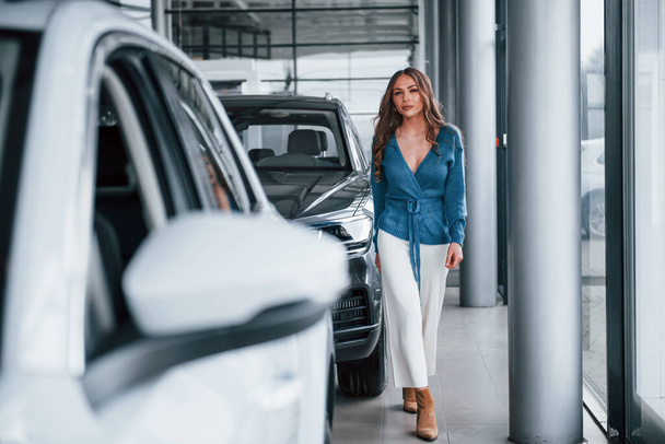 Positive woman in blue shirt walks near brand new car. In auto salon. - Photo, image