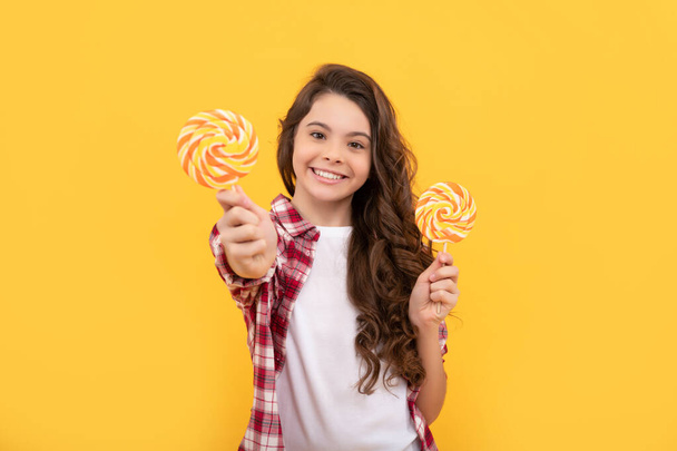 happy child with long curly hair show lollipop caramel candy on yellow background, dental care. - Photo, image