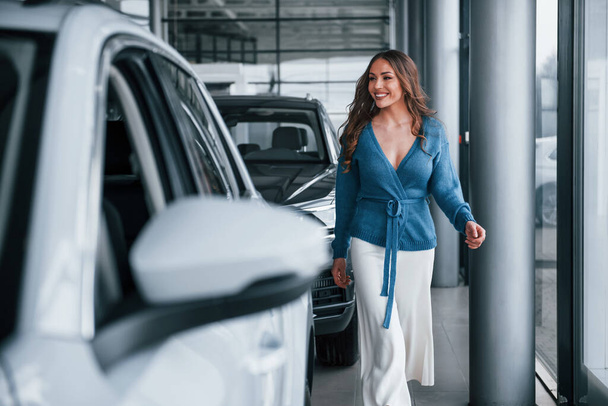Positive woman in blue shirt walks near brand new car. In auto salon. - Фото, зображення