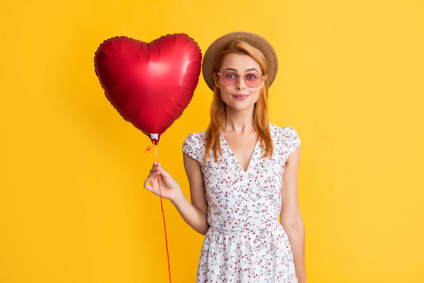 smiling young woman in straw hat and sunglasses hold love heart balloon on yellow background - Zdjęcie, obraz