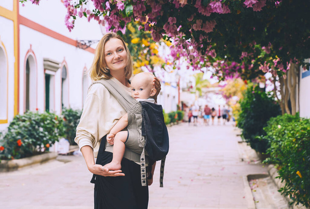Mother and baby on beautiful street full of blooming trees in Puerto Mogan town, Gran Canaria, Spain. Active travel family vacation in Canary Islands. Woman with child journey in Europe. - Foto, Imagem