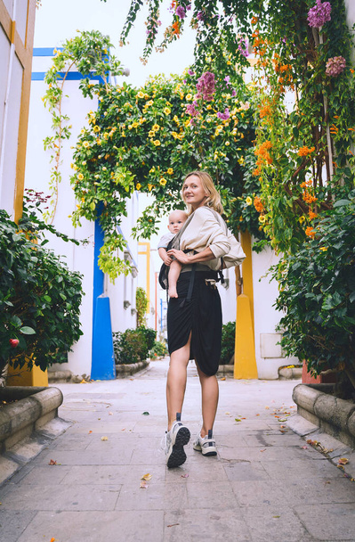 Mother and baby on beautiful street full of blooming trees in Puerto Mogan town, Gran Canaria, Spain. Active travel family vacation in Canary Islands. Woman with child journey in Europe. - 写真・画像