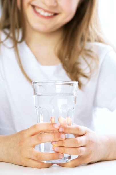 Close-up of a girl in the kitchen drinks clean water from a glass glass. Focus on the glass, face in defocus. The importance of water for children, maintaining water balance - Foto, imagen
