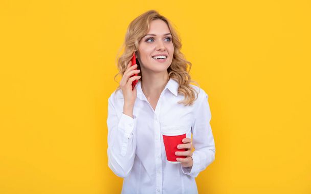 positive blonde woman with coffee cup talking on phone on yellow background - Photo, Image