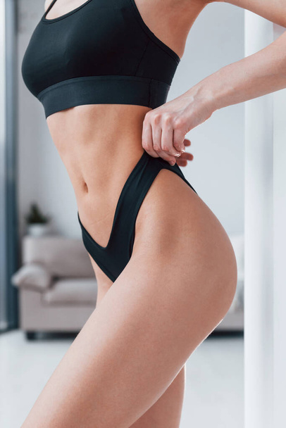 Slender woman in black fitness underwear standing indoors in room at daytime. - Фото, изображение