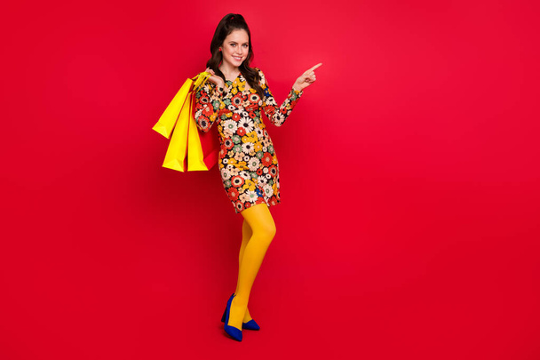 Photo of charming sweet woman wear flower print dress holding shoppers pointing empty space isolated red color background - 写真・画像