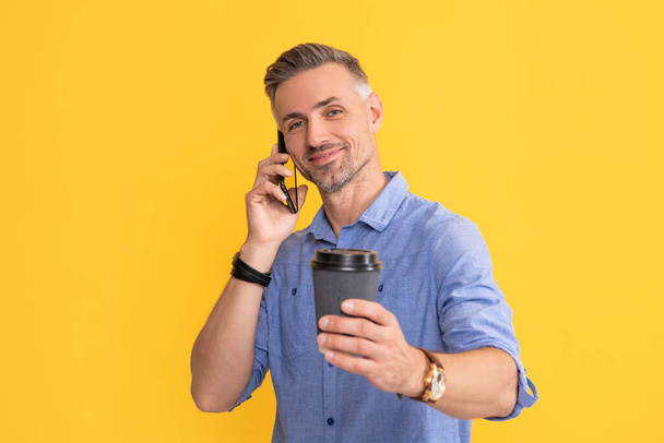 glimlachende man praten over de telefoon met koffie kopje op gele achtergrond, selectieve focus, 's morgens koffie. - Foto, afbeelding