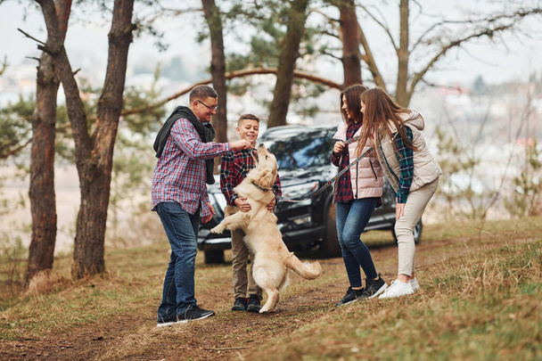 Happy family have fun with their active dog near modern car outdoors in forest. - Photo, Image