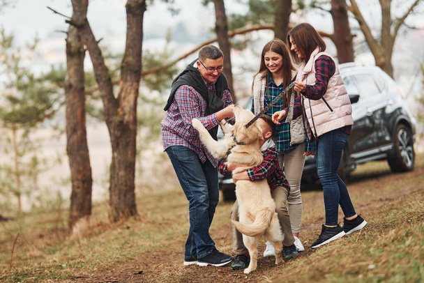 Happy family have fun with their active dog near modern car outdoors in forest. - Photo, image