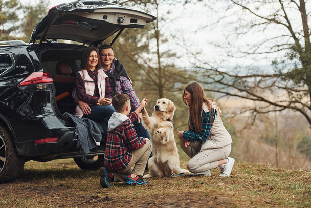 Happy family have fun with their dog near modern car outdoors in forest. - Фото, изображение