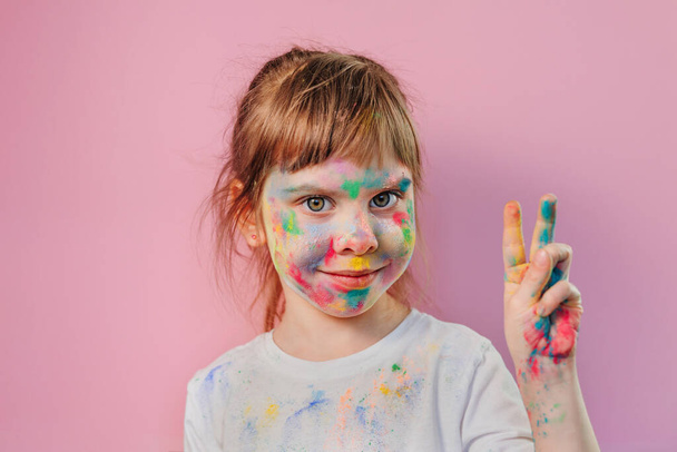 Little girl's face covered with holi colors on pink background. Portrait of happy smiling toddler with colotful paintings on the skin. - Фото, изображение