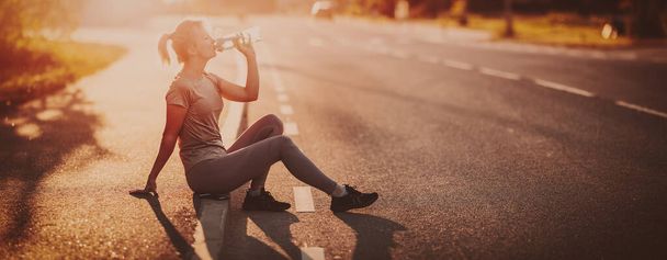 Woman sitting on the border of the roadside after a run and drinking water. - Zdjęcie, obraz