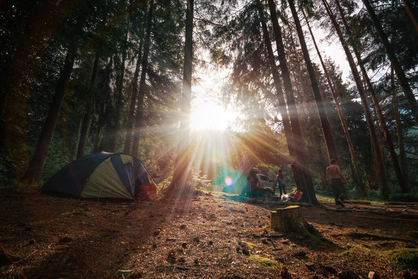 Warm sun rays. Tourist camp among tall pines. Smoke from the fire. Tent. Light rain, drops shining in the sun. People on vacation. Vehicles in the background. Rain cover. - Photo, Image