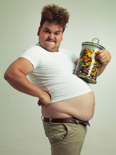 Tohle mi pomůže zvětšit se a zvětšit. Vystřižený snímek muže s nadváhou, který drží velkou sklenici cukroví. - Fotografie, Obrázek