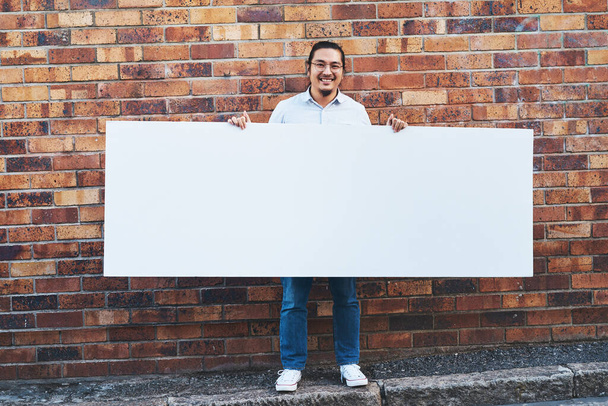 ¿Qué harías con todo este espacio? Fotografía de un joven sosteniendo una pancarta en blanco sobre un fondo urbano al aire libre. - Foto, imagen