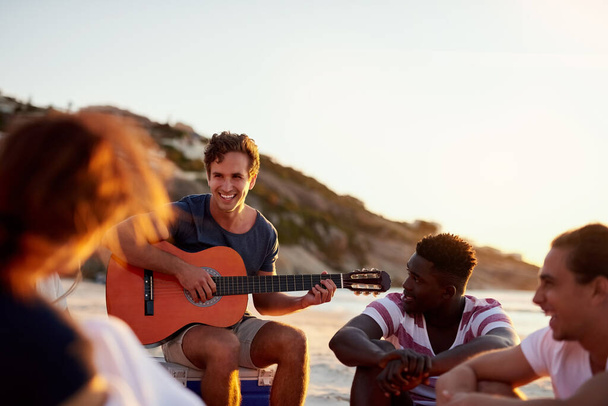 友人は私たちを刺激し、動機づけるものです。友達とビーチに座っている間にギターを弾いている男のショット. - 写真・画像