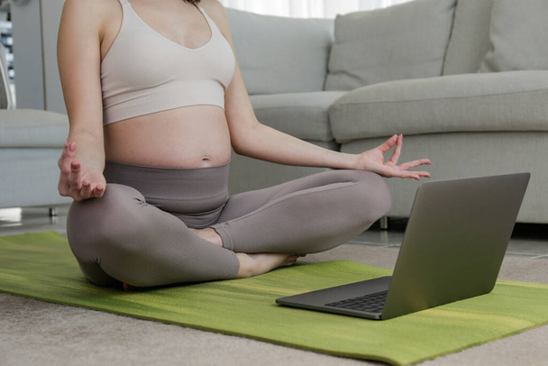 Young beautiful woman on second trimester of pregnancy taking yoga classes online. Pregnant female in sitting on a mat with laptop. Expecting a child concept. Background, copy space, close up. - 写真・画像