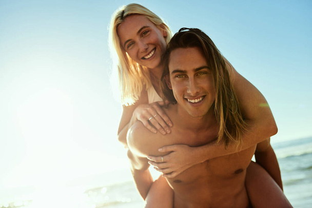 Beach days are a must in summer. Portrait of a young man giving his girlfriend a piggyback ride at the beach. - Photo, Image