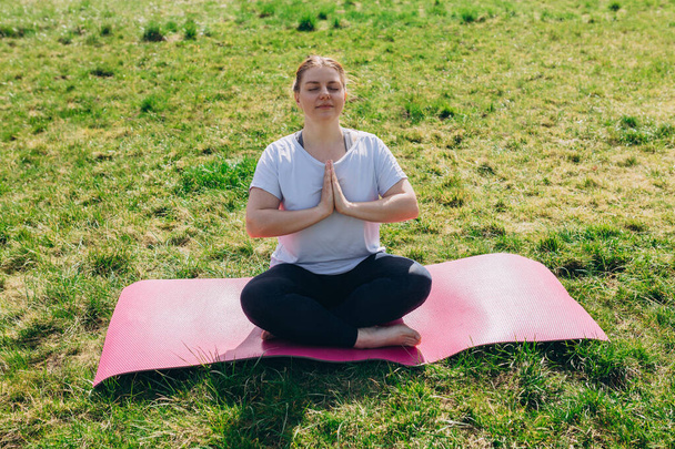 Giovane donna rossa attraente sorridente che pratica yoga, ragazza che fa esercizio Ardha Padmasana e medita in posa Half Lotus con namaste sullo sfondo della natura, lavorando fuori - Foto, immagini