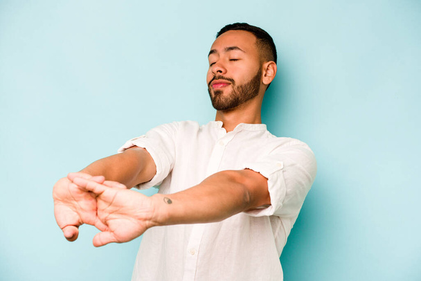 Young hispanic man isolated on blue background stretching arms, relaxed position. - Φωτογραφία, εικόνα