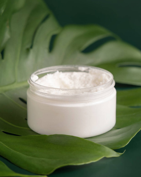 Opened White jar on monstera leaf on green table close up. Brand packaging mockup. Skincare beauty product, cream or butter. Making homemade herbal cosmetics from natural ingredient - Foto, Imagem
