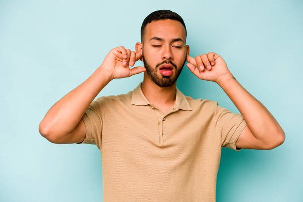 Jonge Spaanse man geïsoleerd op blauwe achtergrond bedekt oren met vingers, gestrest en wanhopig door een luid ambient. - Foto, afbeelding
