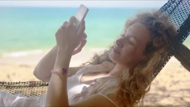 Entspannte Frau surft Smartphone, während sie in der Hängematte am Strand faulenzt - Filmmaterial, Video