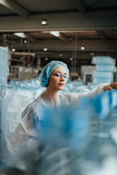 Female worker in protective workwear working in medical supplies research and production factory and checking canisters of distilled water before shipment. Inspection quality control.  - 写真・画像