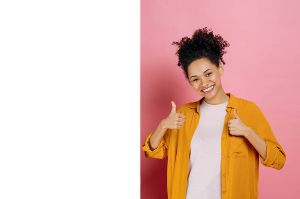 Happy lovely curly african american girl, in casual wear, shows thumb up gesture, stand near empty white space for advertising or presentation, on pink background, looks at camera, smiles. Mock-up - Photo, image
