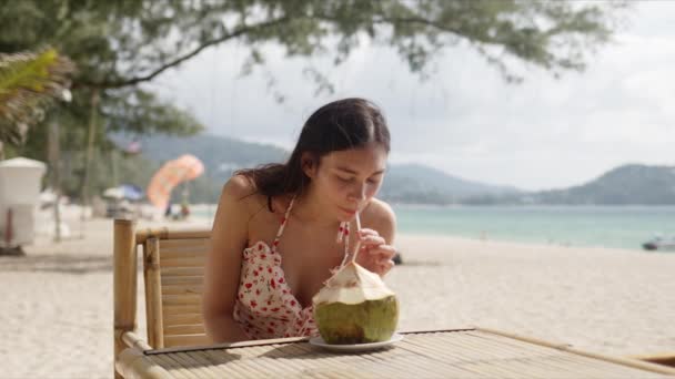 Mladá žena sedí u stolu s kokosem na pláži - Záběry, video