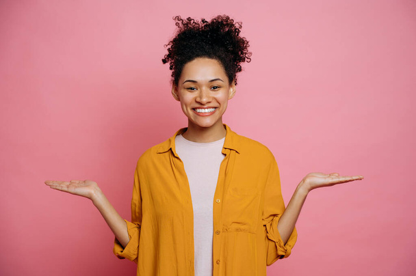 Cheerful african american girl with curly hair in casual clothes, pointing with hands to the sides at an empty space for a presentation, looks at camera, smiling, standing on isolated pink background - Photo, Image