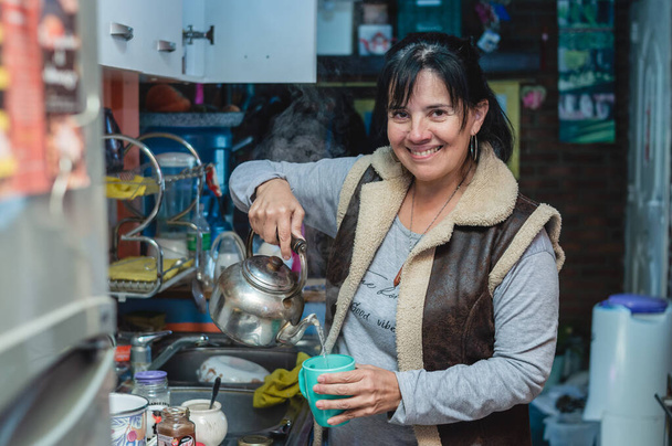 reife kaukasische Argentinierin, die in die Kamera blickt und lächelt, in der Küche ihres Hauses heißes Wasser in einer Tasse serviert und Tee zubereitet. - Foto, Bild