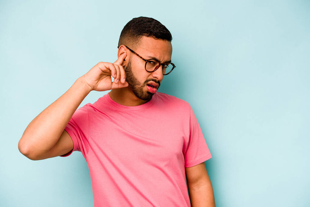 Young hispanic man isolated on blue background covering ears with fingers, stressed and desperate by a loudly ambient. - Photo, Image