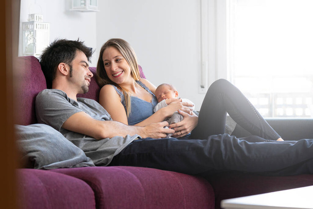 A couple of parents sitting on a sofa in the living room with their newborn baby - Фото, зображення