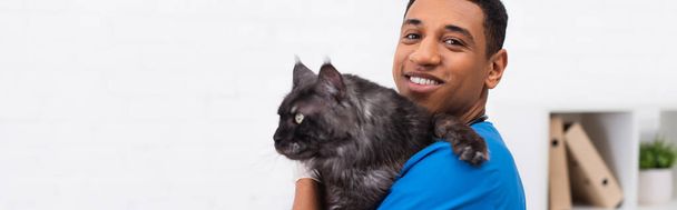 Cheerful african american vet doctor holding maine coon and looking at camera in clinic, banner  - Fotó, kép
