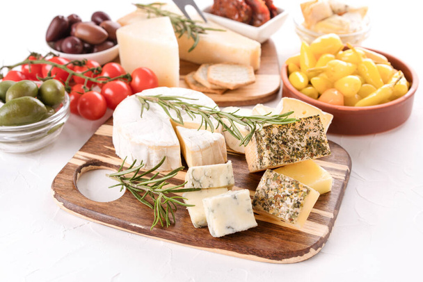 Wooden cheese board with variety of cheeses served with olives, cherry tomatoes, pickled baby peppers on white table background. Cheese plate for aperitif - brie, dorblu, gorgonzola, camembert and parmesan - Foto, imagen