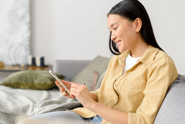 Side view of the calm asian woman sitting on the couch in cozy living room and holding smartphone. Female spending time online, texting with friends - Fotoğraf, Görsel